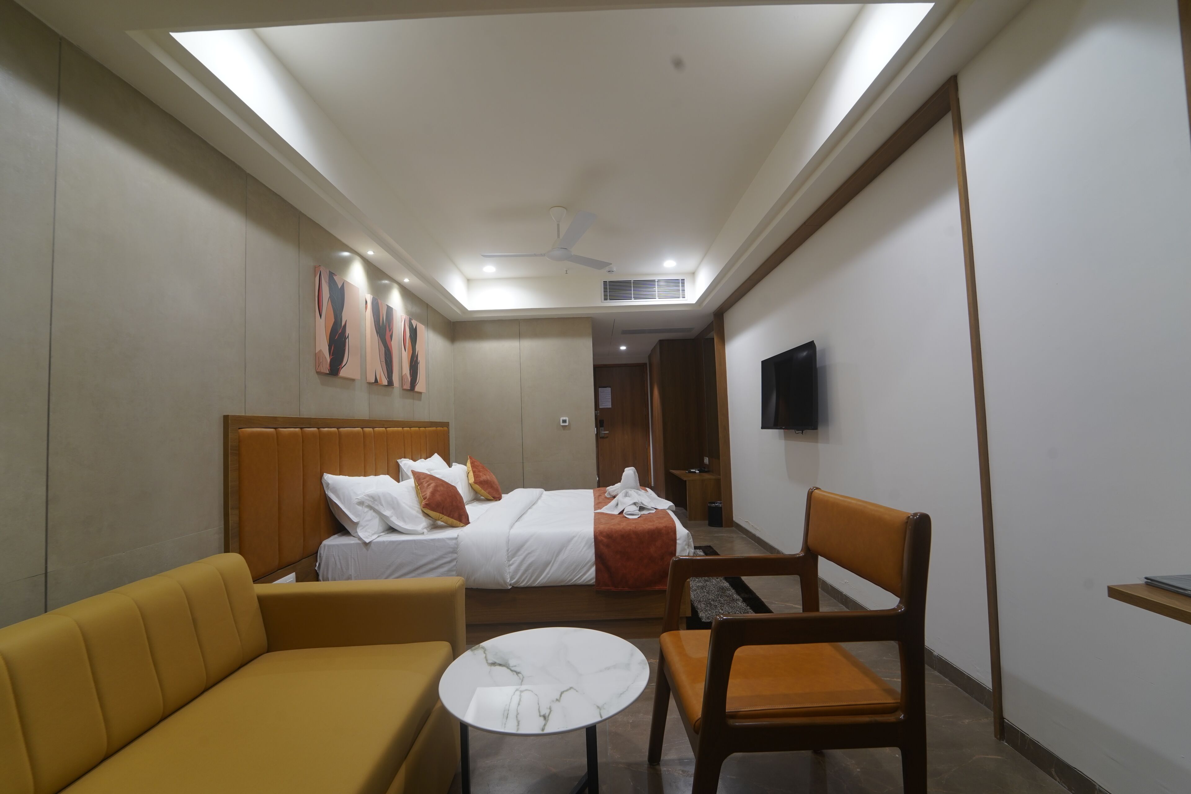 A hotel room with a bed, a sofa, a chair, a small table, a TV mounted on the wall, and a ceiling fan.