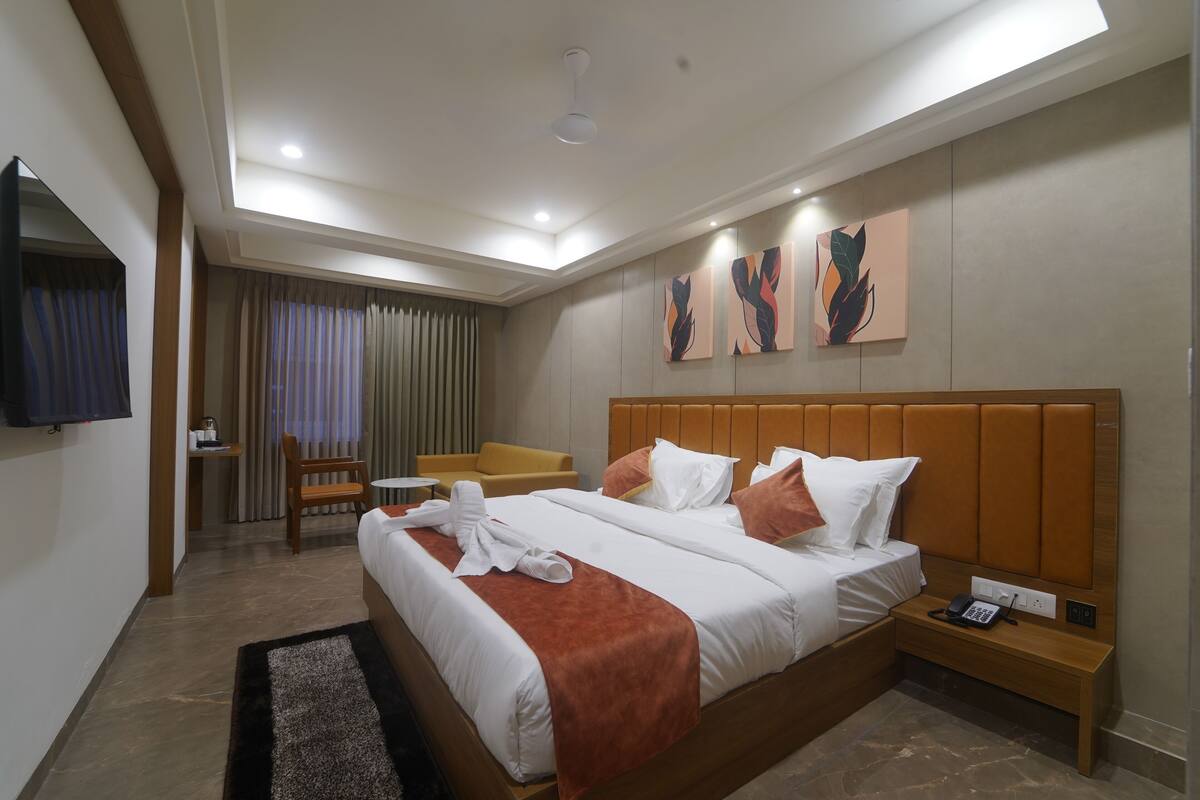 A hotel room with a large bed, a bedside table, a chair, a desk, and a television on the wall.