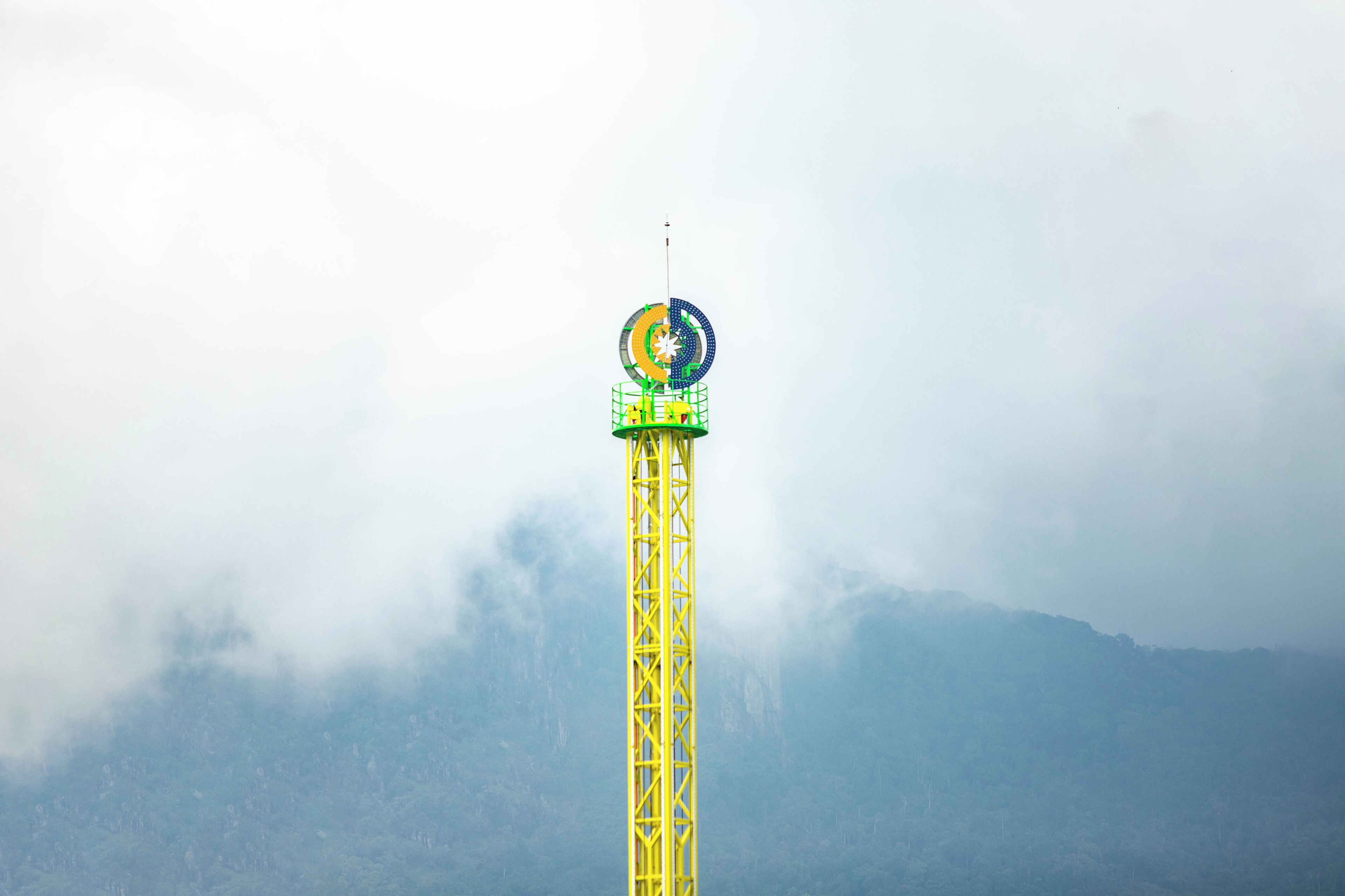 A tall yellow tower with a green platform and a circular structure on top, surrounded by fog and mountains.