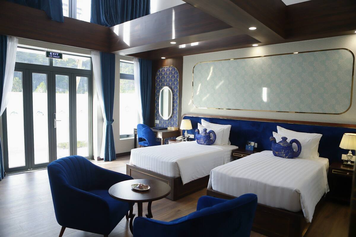Une chambre d’hôtel avec deux lits, une petite table, deux chaises, un miroir et une grande fenêtre.