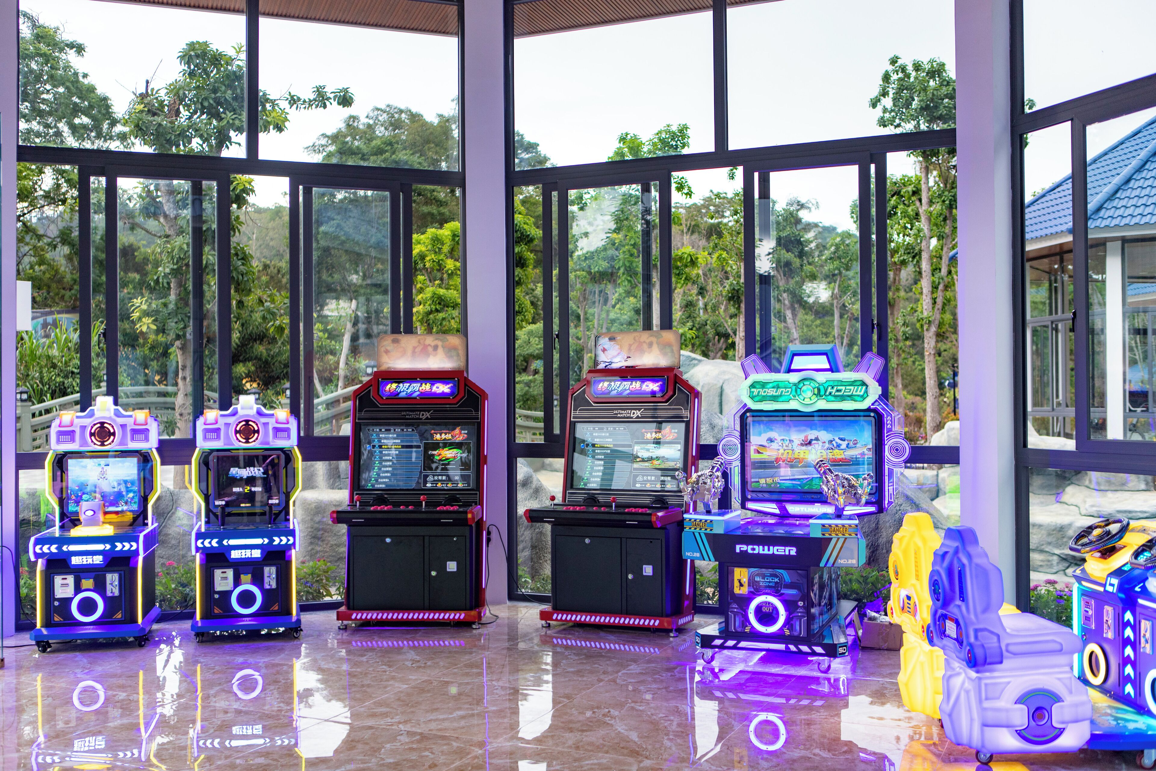 Une rangée de bornes d’arcade dans une pièce spacieuse et bien éclairée, dotée de grandes fenêtres qui offrent une vue sur les arbres et un bâtiment à l’extérieur.