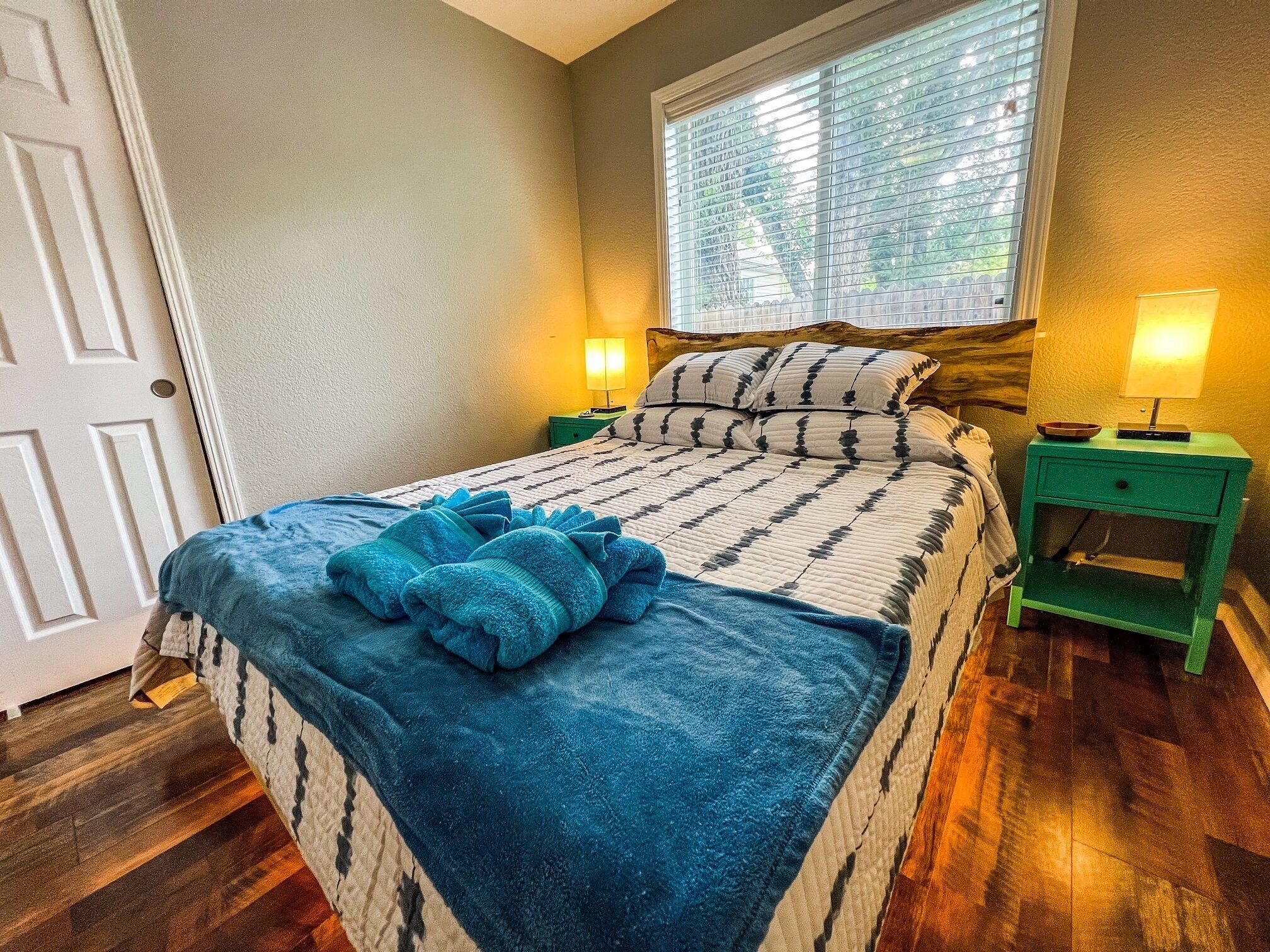 Uma cama bem arrumada com um cobertor azul, duas mesas de cabeceira com abajures, uma janela com persianas e um piso de madeira.