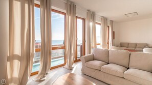 Uma sala de estar moderna com sofá bege, janelas amplas com cortinas e vista para o mar.