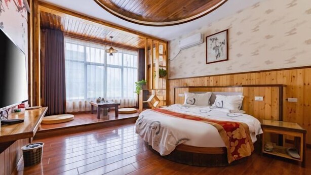 A hotel room with a wooden floor, a bed with a red and white bedspread, a wooden headboard, a small bedside table, a TV, and a window with curtains. - Mount Hua Hongchen Xiake Themed Homestay (Huayin)