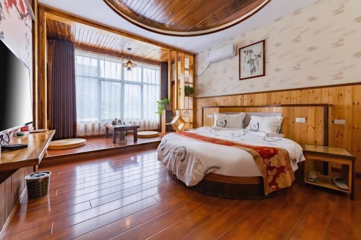 A hotel room with a wooden floor, a bed with a red and white bedspread, a wooden headboard, a small bedside table, a TV, and a window with curtains.