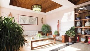 A room with a wooden ceiling, a glass coffee table, a sofa, a bookshelf with decorative items, and a large potted plant. - Mount Hua Hongchen Xiake Themed Homestay (Huayin)