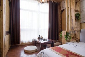 Una habitación con una cama, un otoman redond, una mesa de madera con una tetera y una planta.