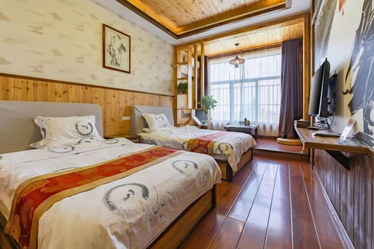 A hotel room with two beds, wooden paneling, a large window with curtains, a TV, and a plant.