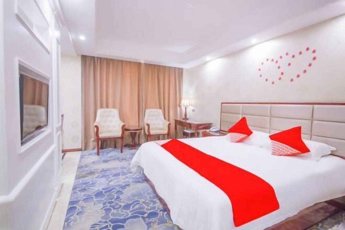 A hotel room with a bed, two chairs, a small table, a TV, and a heart-shaped decoration on the wall.