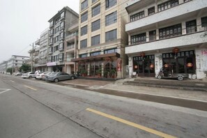 A multi-story building with commercial establishments on the ground floor, a parking area with cars, and a wet street. - Wenzhou Dongtou Overseas Chinese Hotel (Wenzhou)