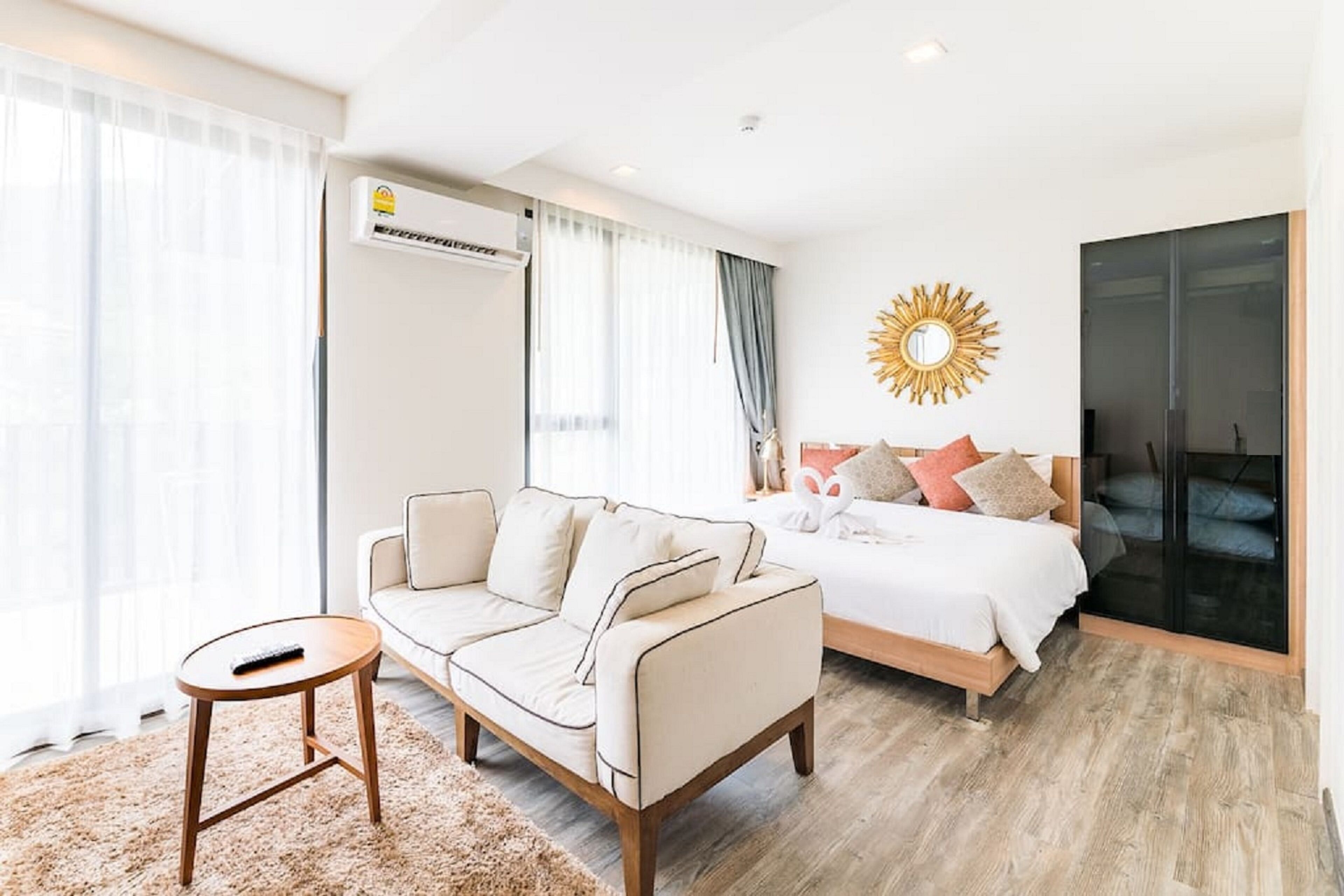A modern hotel room with a sofa, a small round table, a bed with pillows, and a large mirror on the wall.