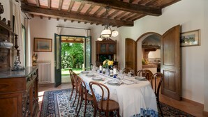 Comedor con una mesa dispuesta para seis comensales, sillas de madera, una araña y vistas del jardín a través de una puerta abierta.