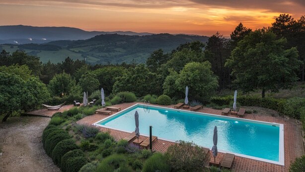 Ein rechteckiger Pool mit einer Ziegelterrasse, umgeben von üppiger Vegetation und Bäumen, mit einem malerischen Ausblick auf entfernte Hügel und einem Sonnenuntergang im Hintergrund.
