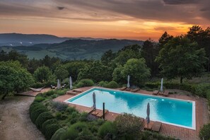 Une piscine rectangulaire bordée d’une terrasse en briques, entourée d’une végétation luxuriante et d’arbres, offrant une vue pittoresque sur des collines lointaines et un coucher de soleil en arrière-plan.