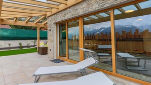 A modern outdoor lounge area with wooden beams and stone walls, featuring a glass door leading to a dining area with tables and chairs. - Accommodation with sauna near the ski lift (Mittersill)