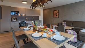 A modern dining area with a wooden table set for a meal, a kitchen in the background, and a gray couch. - Accommodation with sauna near the ski lift (Mittersill)