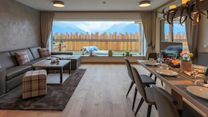 A modern hotel room with a sofa, dining table, and a view of a wooden fence and mountains.