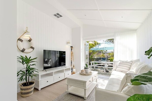 A modern living room with a flat-screen TV, white furniture, a large window with a view of the outdoors, and a ceiling fan.