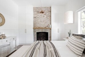 A bedroom with a brick fireplace, white cabinetry, a bed with striped bedding, and a window providing natural light.