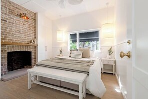A bedroom with a brick fireplace, a bed with a bench, bedside tables, and a window with curtains. - Steamers Bungalow (Huskisson)