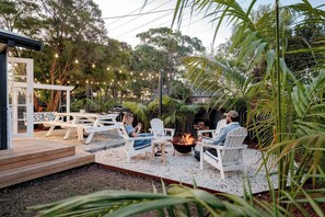 An outdoor patio with white Adirondack chairs, a fire pit, and a wooden deck. - Steamers Bungalow (Huskisson)