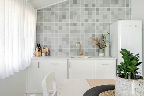 A modern kitchen with white cabinets, a grey tiled backsplash, a gold faucet, and a white refrigerator.