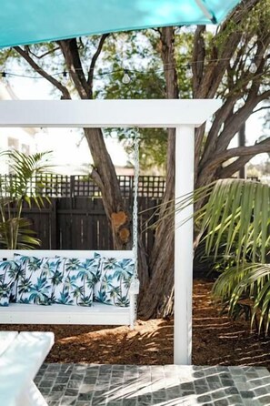 A covered outdoor seating area with a swing, surrounded by trees and a fence.