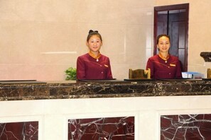 Two receptionists at a hotel reception desk with marble panels and a wooden door in the background. - Shenhang Yinju Mingzhu Hotel (Jiashan)