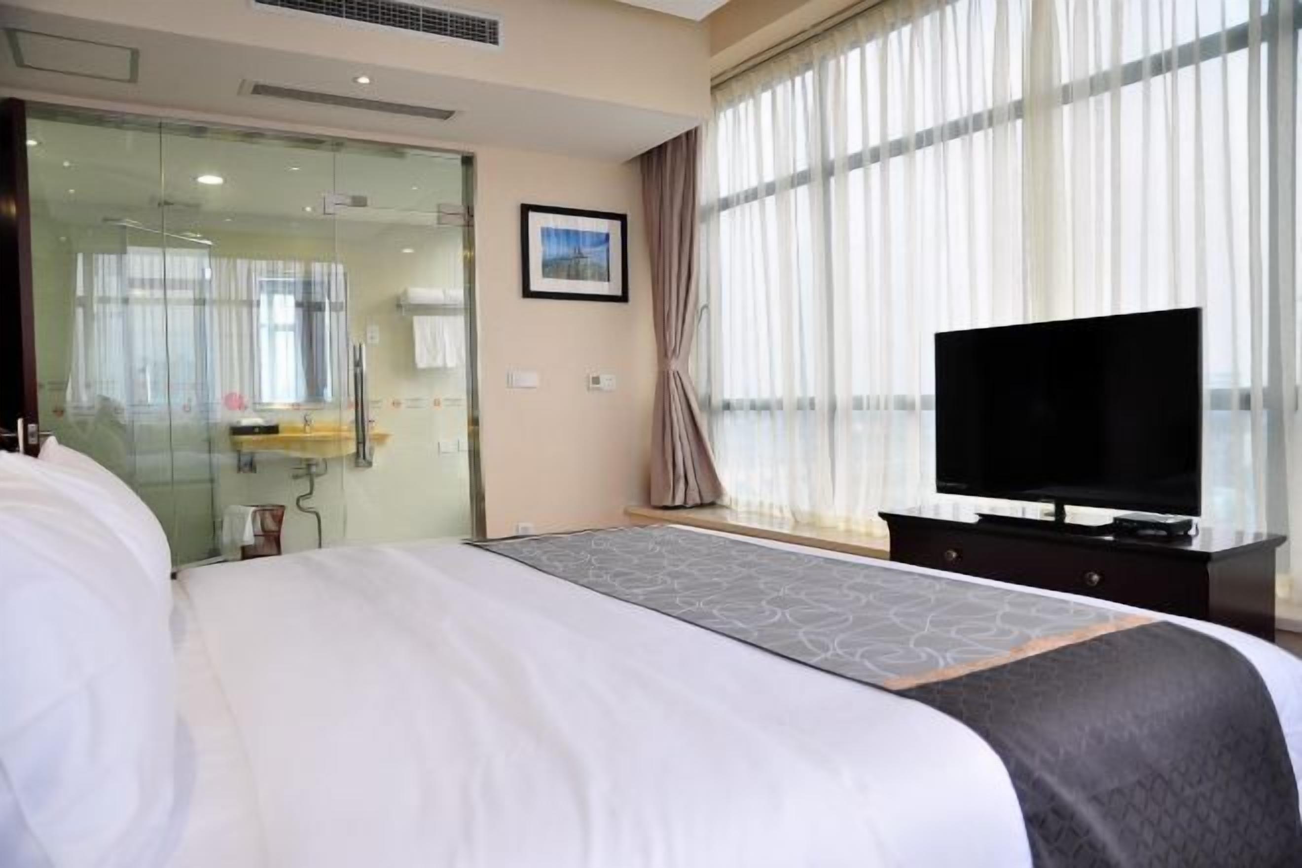 A hotel room with a large bed, a television, a glass partition, and a window with curtains.