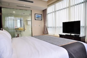 Quarto de hotel com cama grande, televisão, divisória de vidro e janela com cortinas.