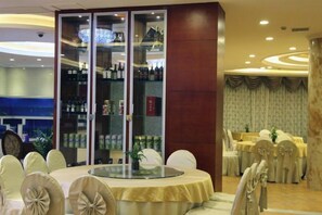 Espaço de refeições de hotel com mesas redondas, cadeiras e uma vitrine de vidro com garrafas.