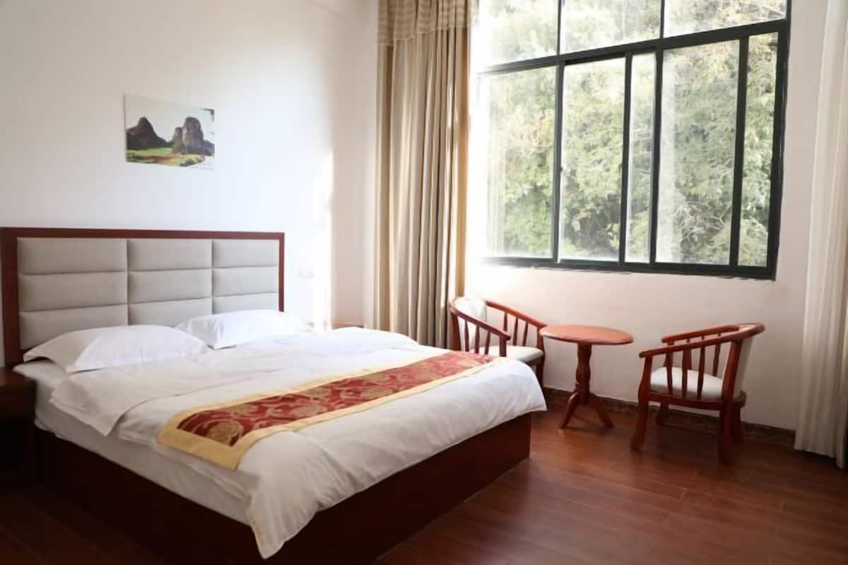 A bedroom with a bed, a small table, two chairs, a window with curtains, and a painting on the wall.