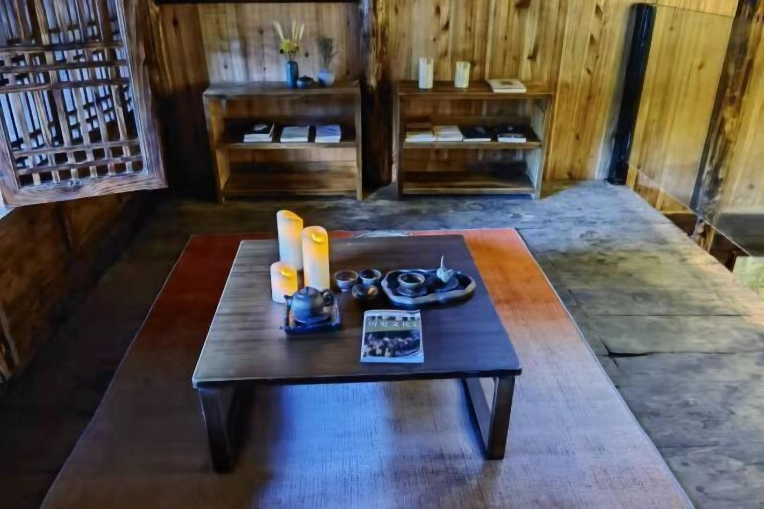 Ein Holzinterieur mit einem Tisch, der mit Kerzen, einem Teekessel und einer kleinen Ablageplatte gedeckt ist. Im Hintergrund sind Regale mit Büchern und Dekorationen zu sehen.