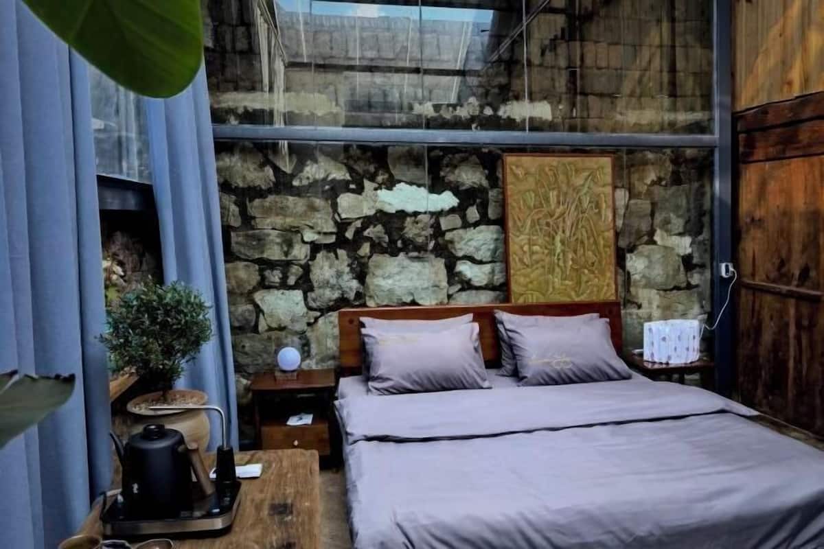 A bedroom with a stone wall, a bed with grey bedding, a wooden headboard, a nightstand with a lamp, and a small table with a kettle.