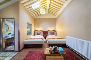 Una camera d'albergo con due letti, un tappeto rosso, un tavolino da caffè in legno con vari oggetti, un soffitto con travi in legno e un termosifone.