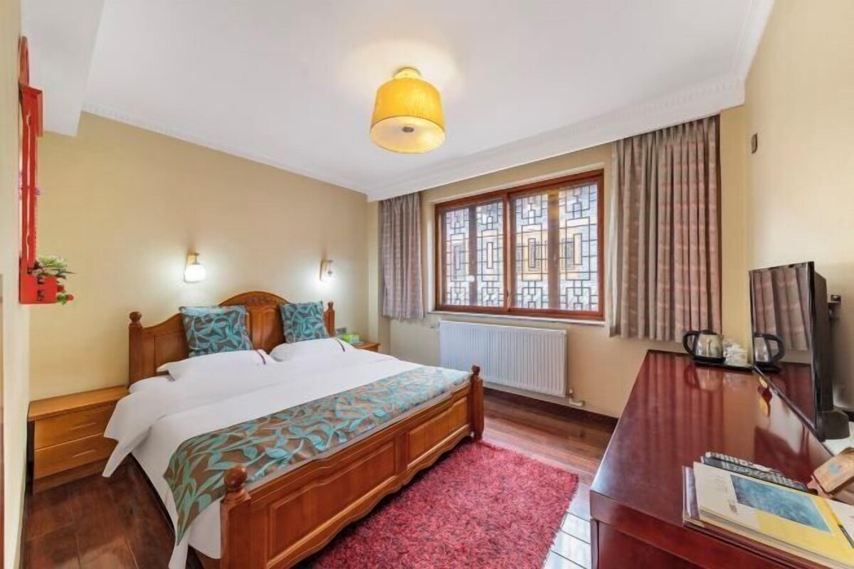 A bedroom with a wooden bed, a red rug, a desk with a chair, a television, and a window with curtains.