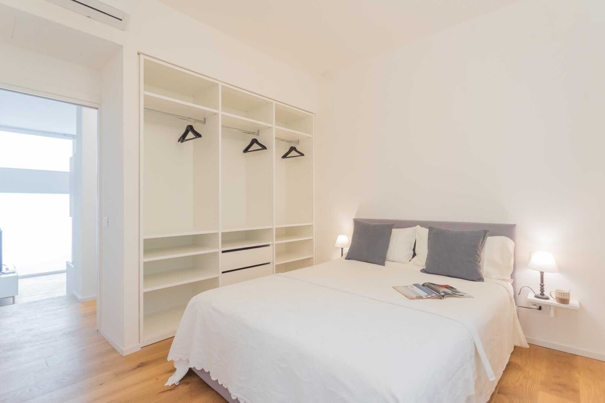 A bedroom with a large white bed, a grey headboard, and a white wardrobe with multiple shelves and hangers.