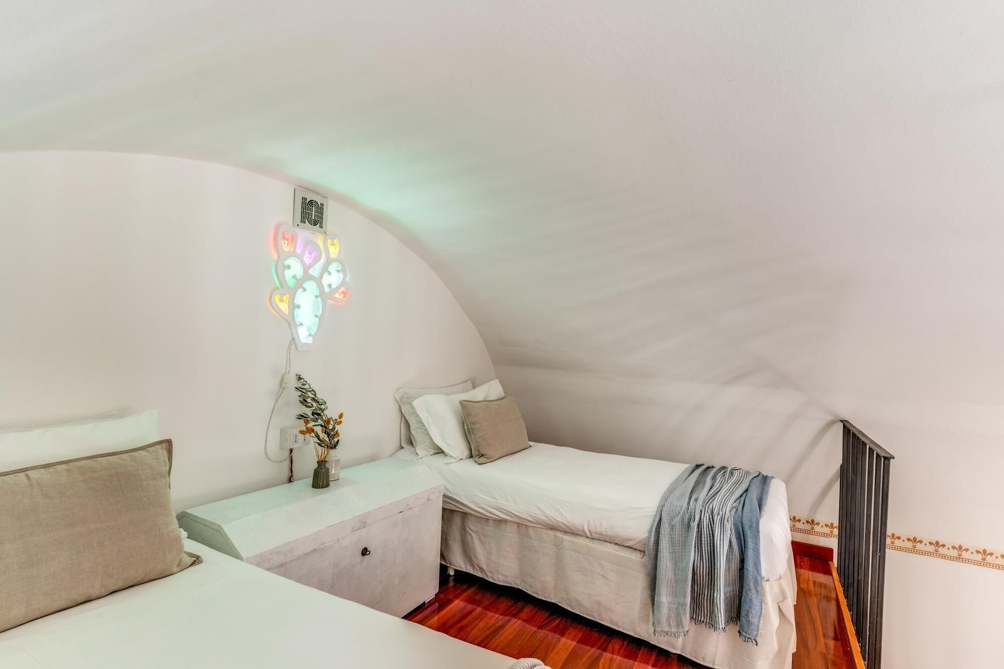 A cozy, compact room with a curved ceiling, a bed with a striped blanket, a bedside table with a vase, and a wall-mounted light fixture.