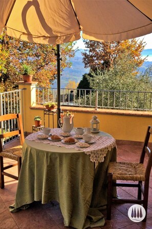 A round table with a lace tablecloth, surrounded by wicker chairs, set outdoors under a large umbrella. - Vacation home nestled in the Tuscan countryside with a private pool (Gallicano)