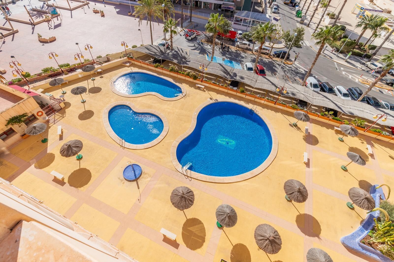 An aerial view of a resort with multiple swimming pools, sun umbrellas, and palm trees.