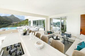 A modern, well-lit living area with a large window overlooking a pool and mountains. There are lounge chairs, a dining table with white chairs, and a sofa with cushions. - Numa Indigo BAY (INDIGO BAY)
