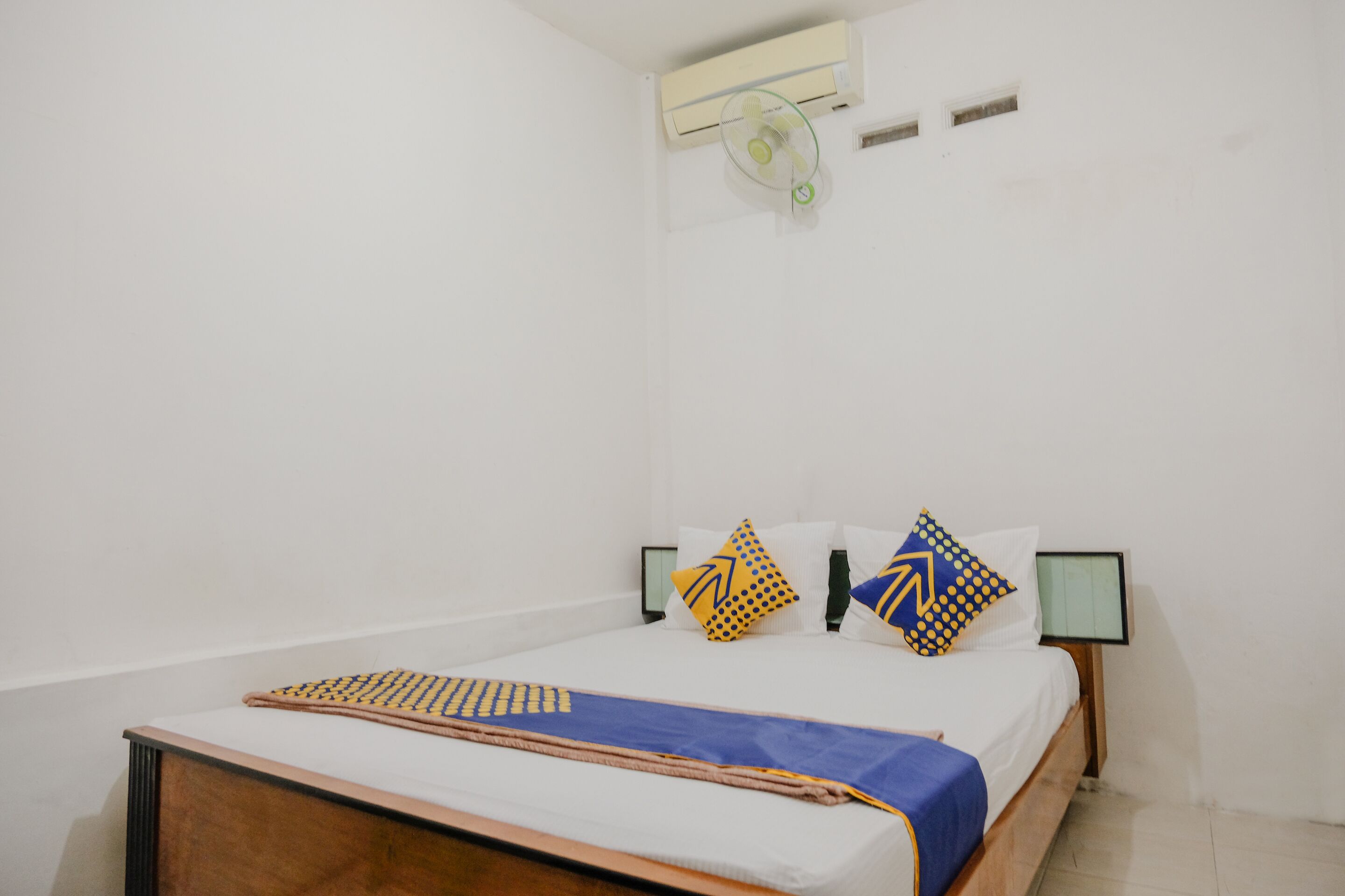 Un lit bien fait, recouvert d’une couvre-lit bleue et beige, avec deux oreillers aux motifs géométriques, et un climatiseur fixé au mur.