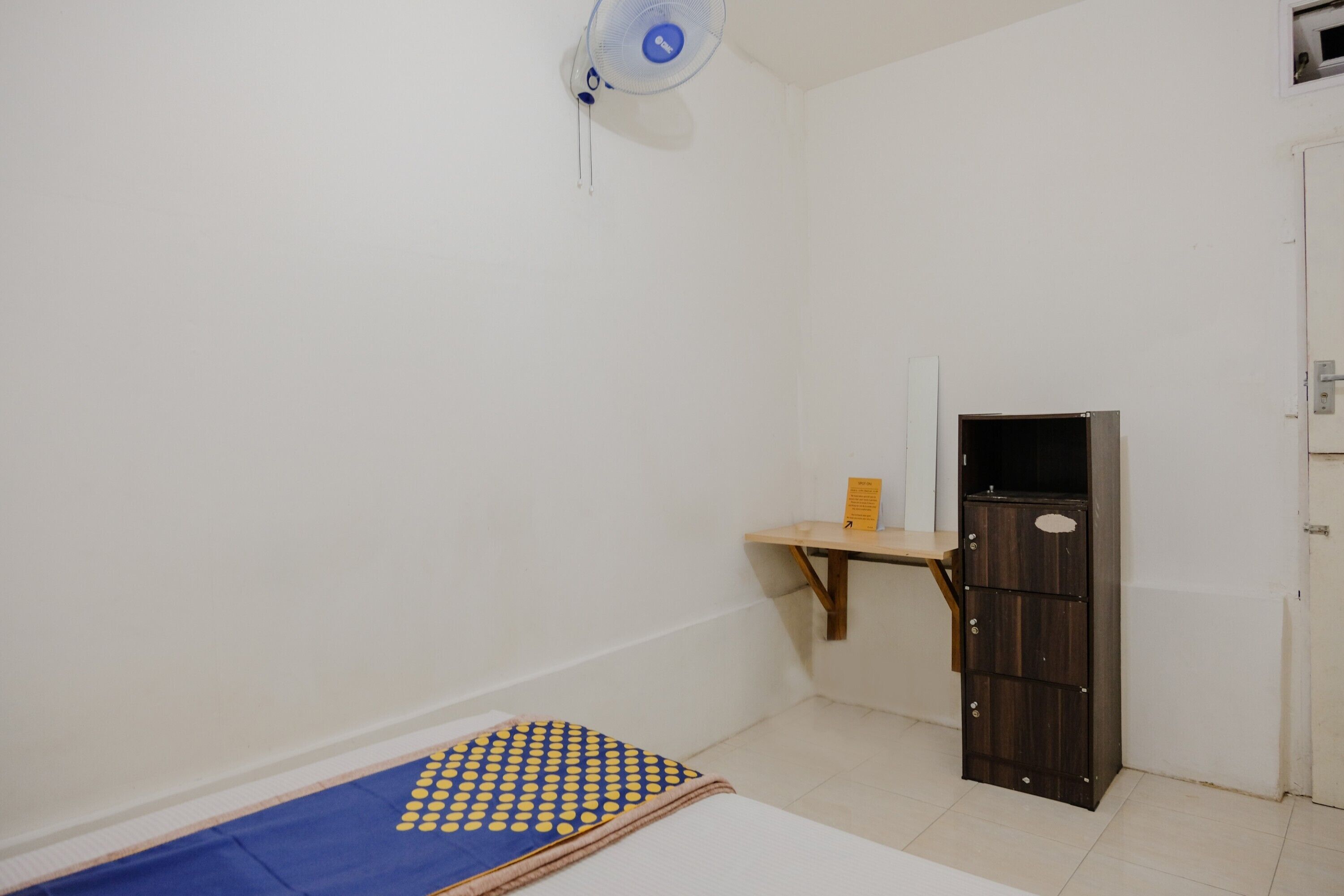 A small, sparsely furnished room with a bed, a small table, a cabinet, and a wall-mounted fan.