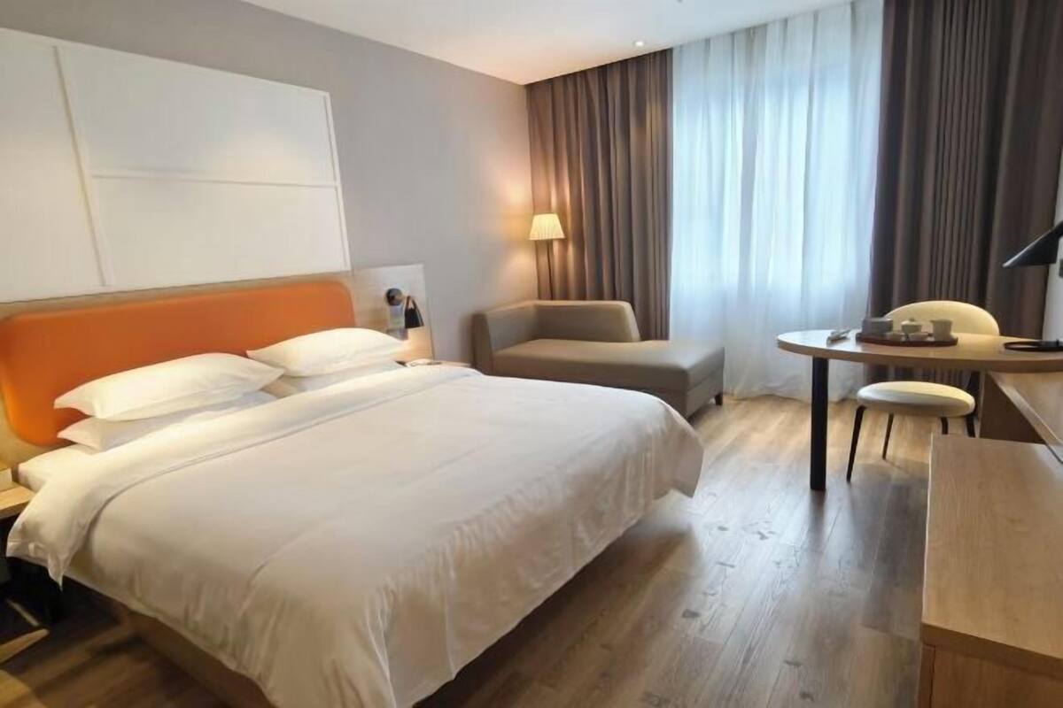 A hotel room with a bed, a desk, a chair, a lamp, and a window with curtains.