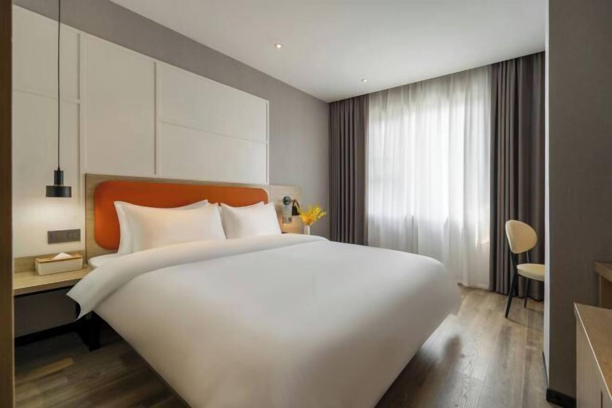 A modern hotel room with a large bed, a bedside table, a nightstand, a chair, and a window with curtains.