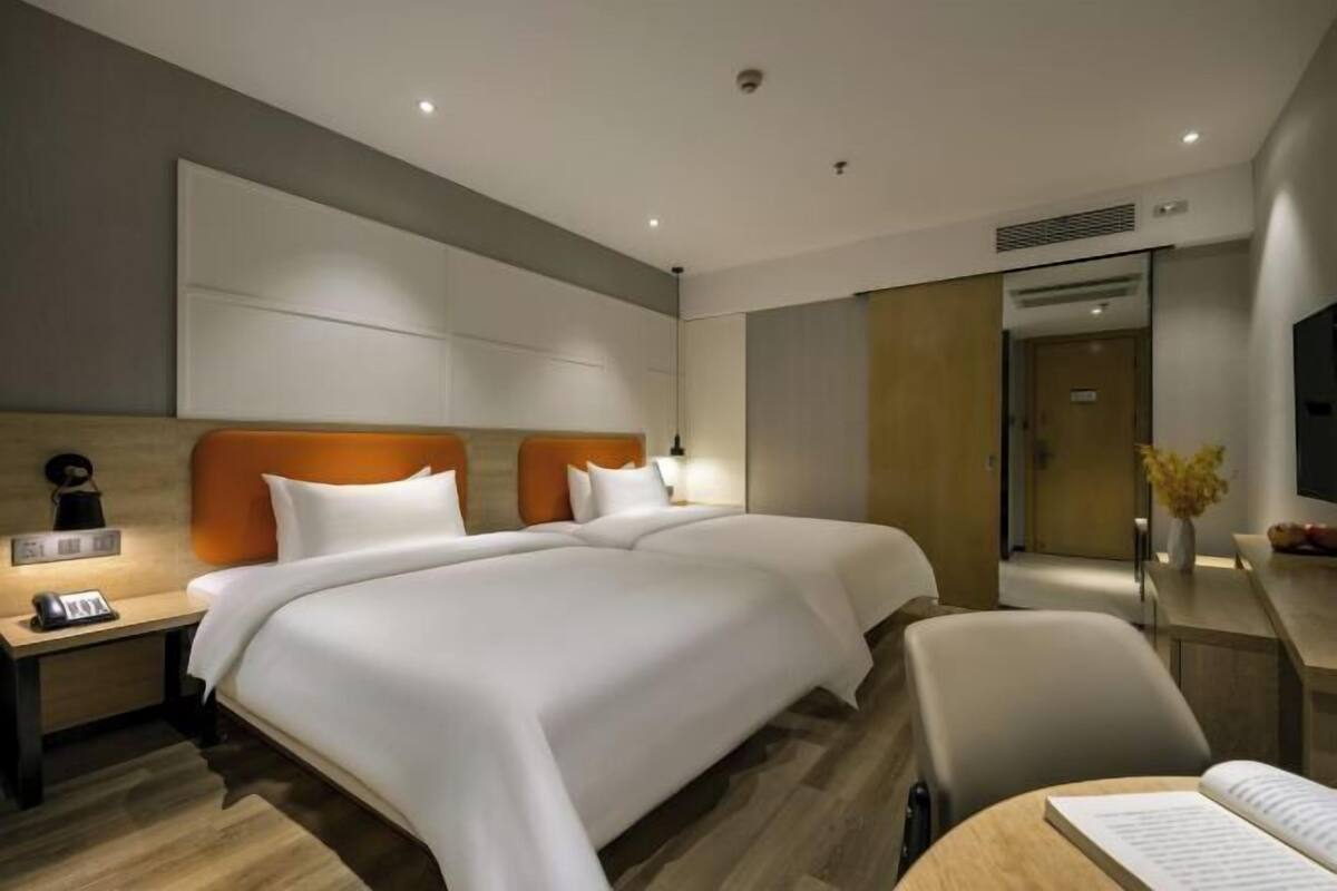 A hotel room with two beds, a desk, a chair, and a small table with a book.