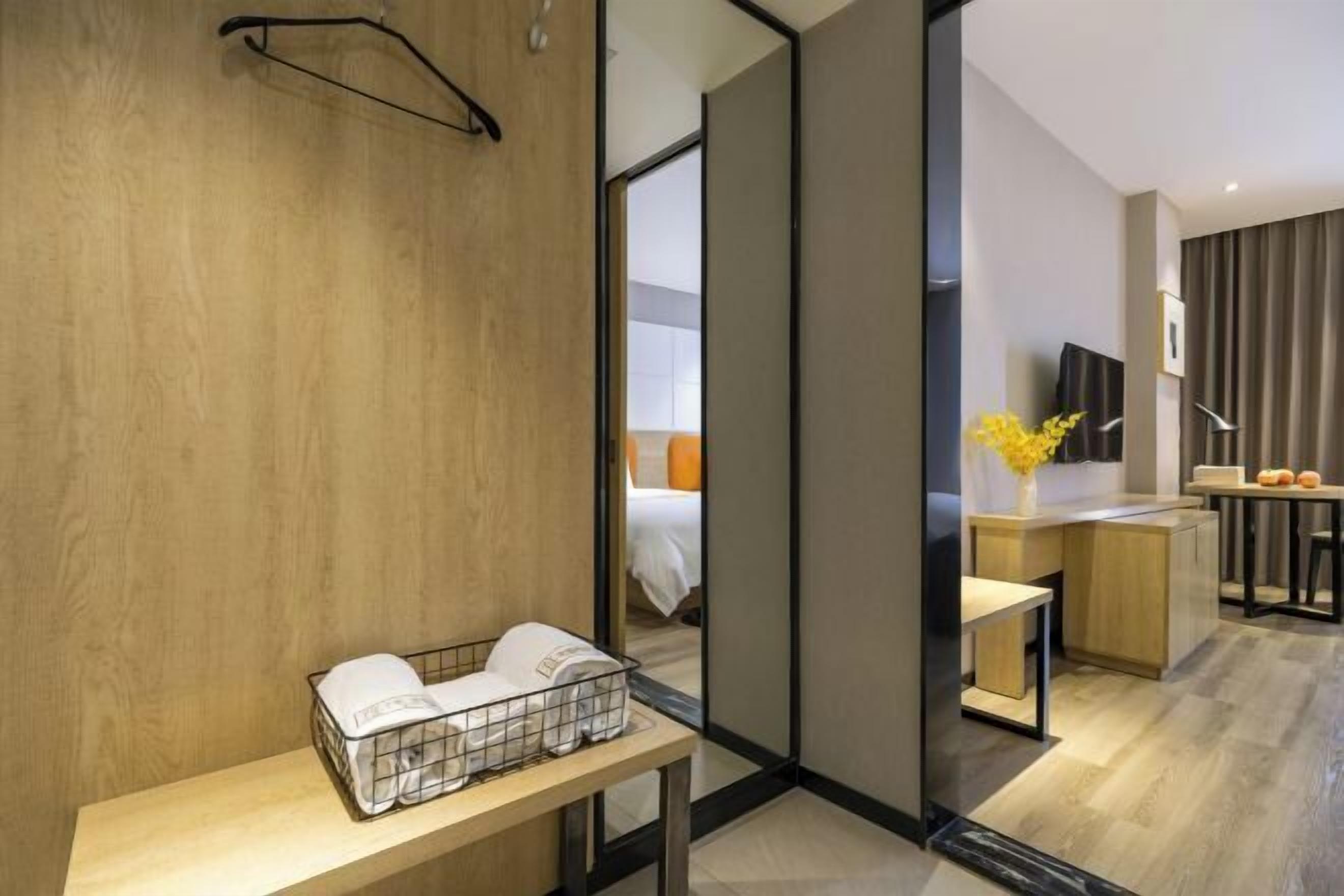 Une chambre d’hôtel moderne avec un mur en bois, une petite table avec des serviettes, un miroir, un bureau avec une chaise et une vue sur la chambre adjacente.