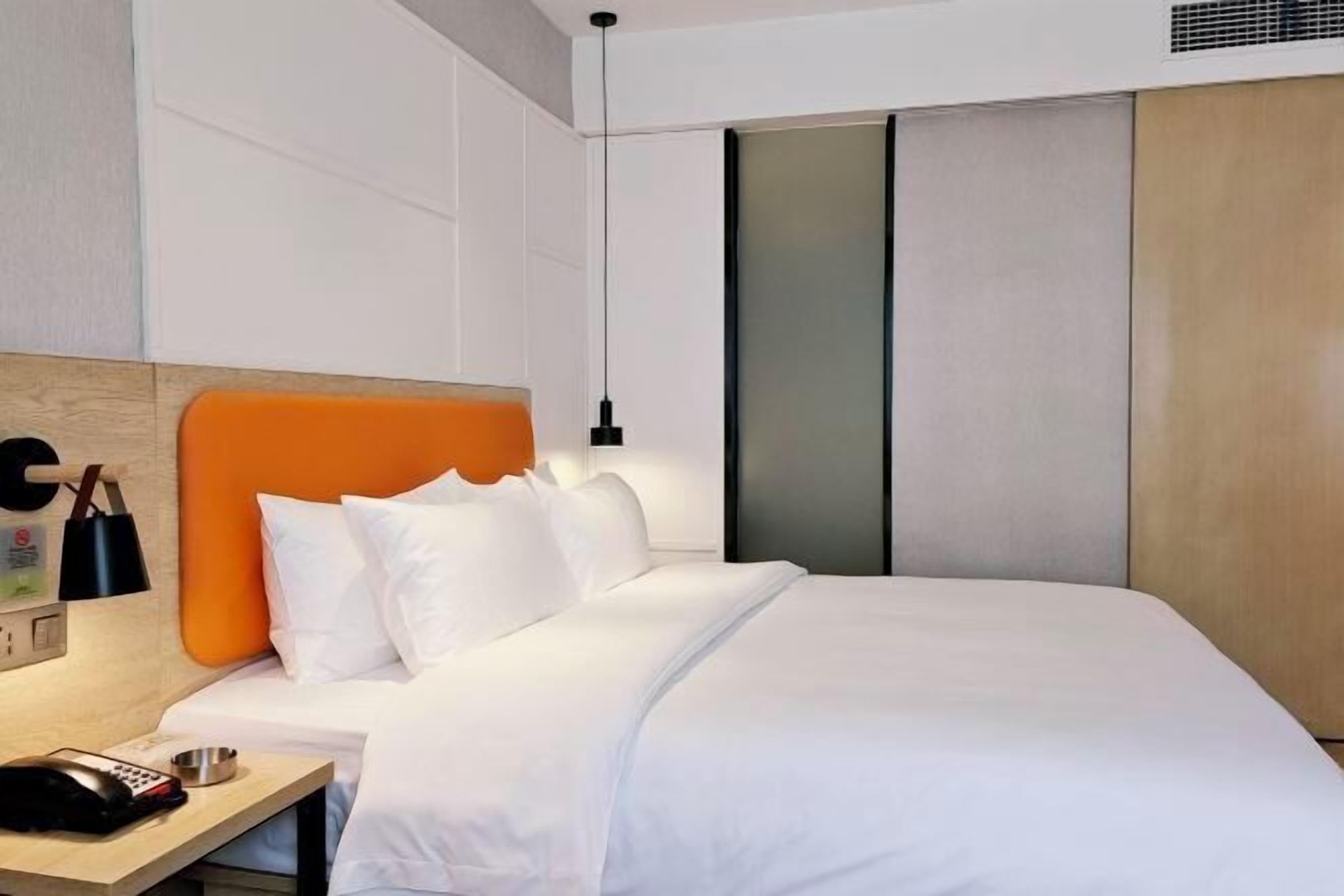 A modern hotel room with a wooden headboard, white bedding, a bedside table with a phone, and a wall-mounted light.