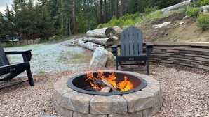 A stone fire pit with a fire burning, surrounded by a stone wall and gravel. There are two Adirondack chairs and a bench nearby. - River's Bend Lodge- Hot Tub, Fire Pit, Games & Ski (Leavenworth)