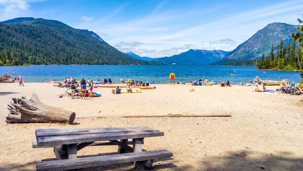 A beach with a wooden picnic table, people relaxing, and mountains in the background. - River's Bend Lodge- Hot Tub, Fire Pit, Games & Ski (Leavenworth)